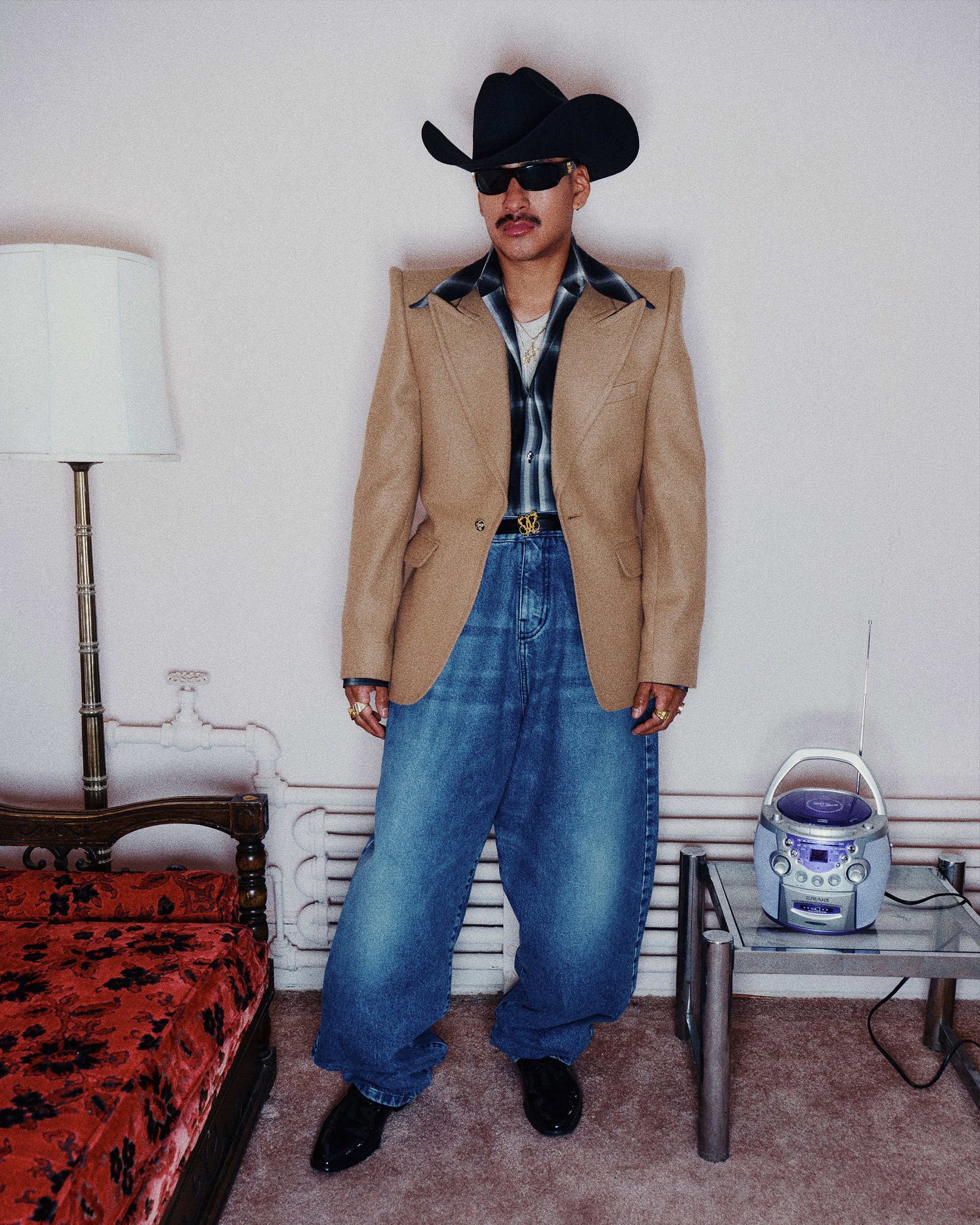 Man wearing a brown blazer, cowboy hat, and baggy jeans standing indoors by a vintage lamp and boombox.