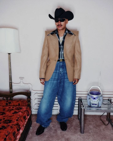 Man wearing a brown blazer, cowboy hat, and baggy jeans standing indoors by a vintage lamp and boombox.