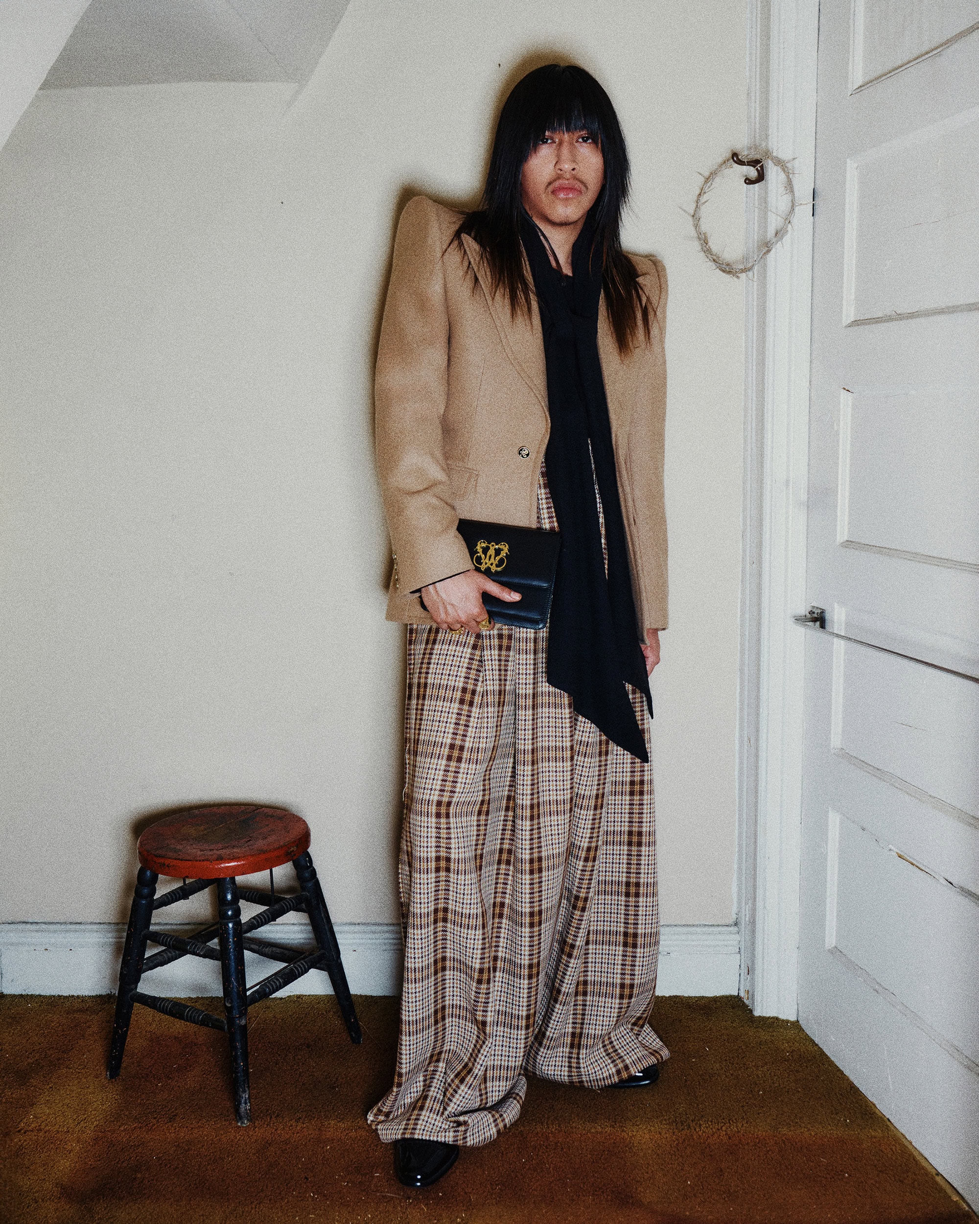 Model standing indoors wearing a structured tan blazer, patterned trousers, and holding a black clutch bag.