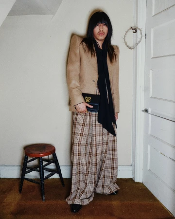 Model standing indoors wearing a structured tan blazer, patterned trousers, and holding a black clutch bag.