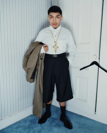 Young man in a white button-down shirt and black shorts holding a patterned coat against a paneled door