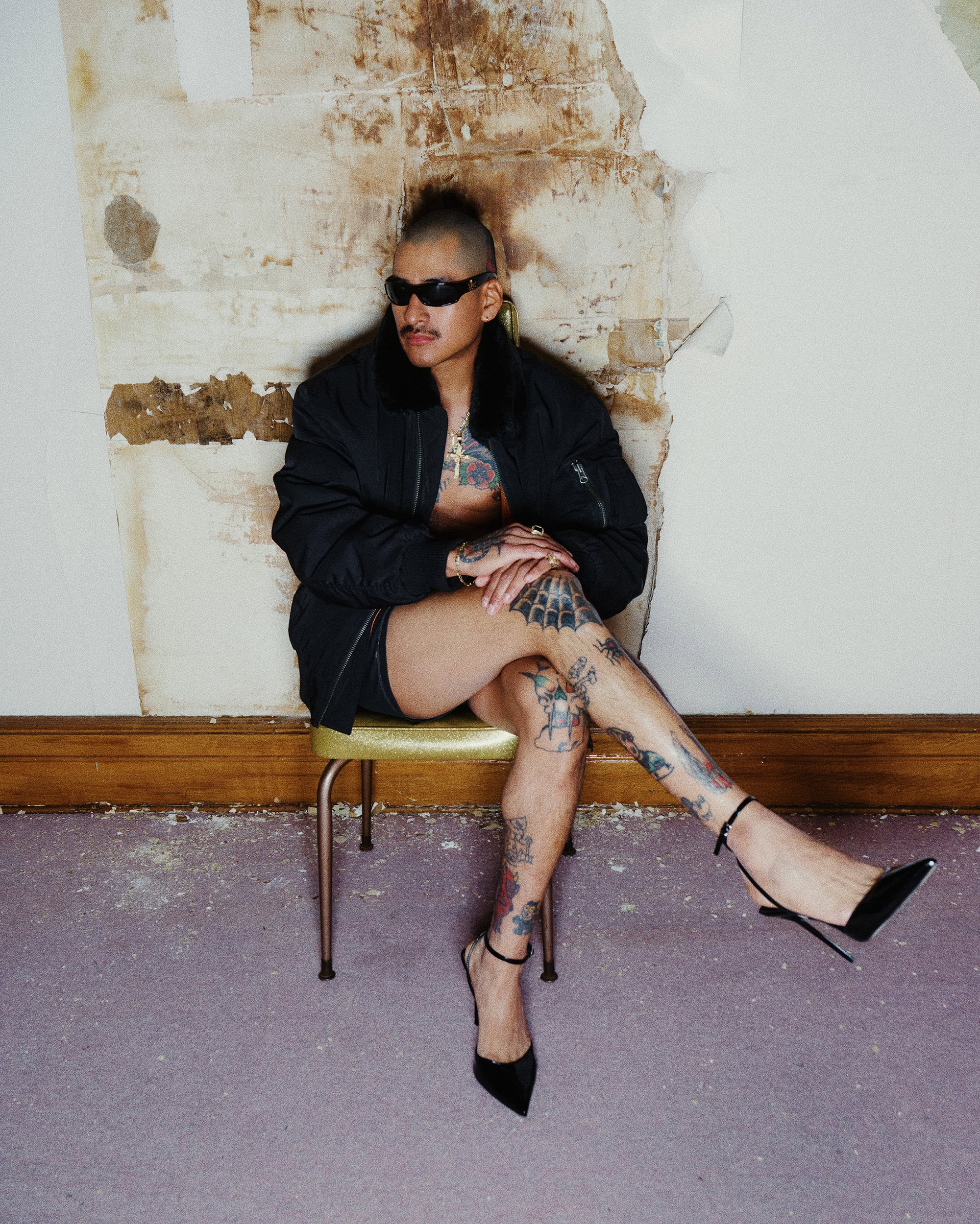 Person with tattoos wearing a black bomber jacket and high heels sitting on a chair against a textured wall.