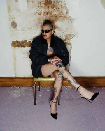 Person with tattoos wearing a black bomber jacket and high heels sitting on a chair against a textured wall.