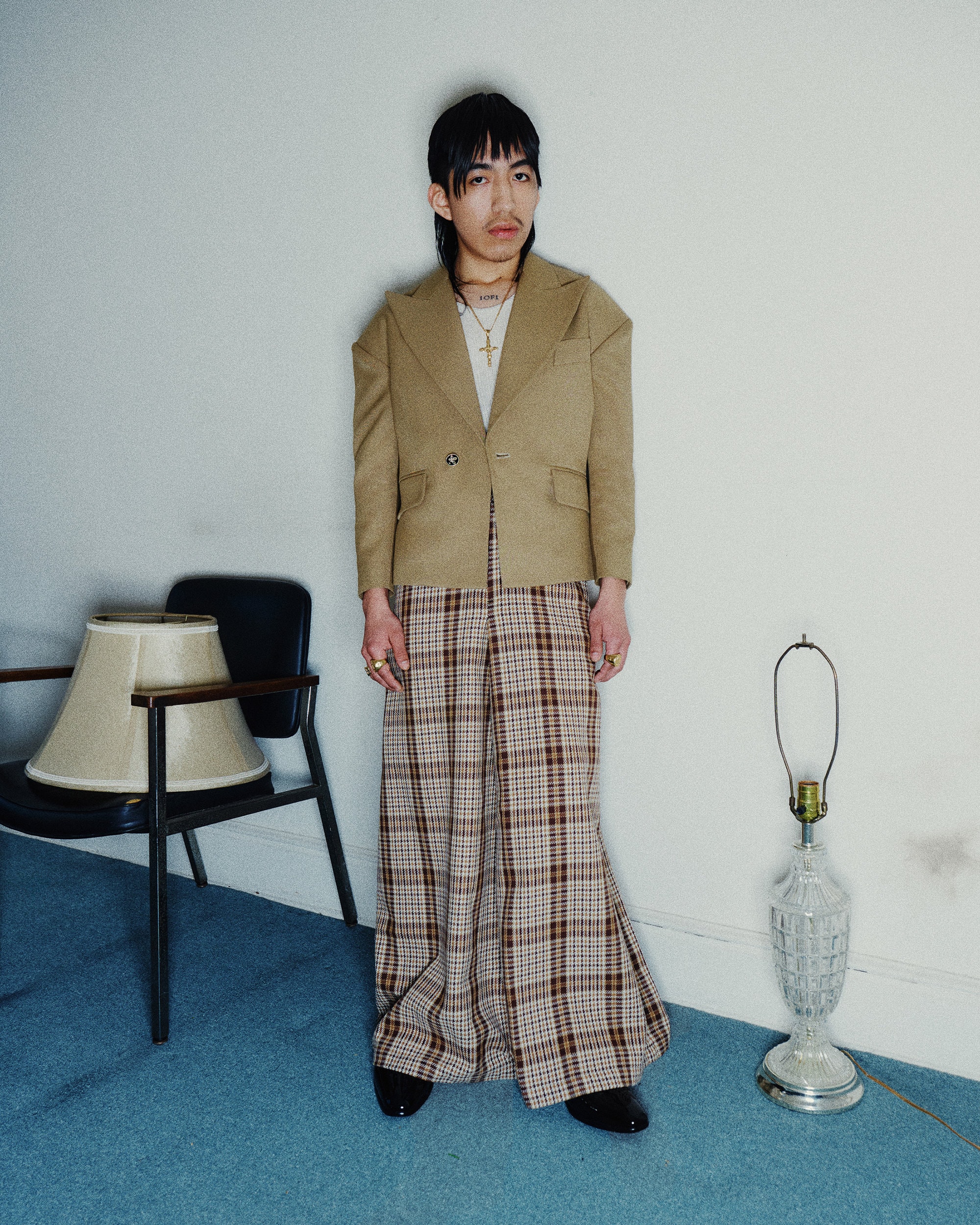 Man wearing a structured tan blazer and plaid trousers standing in an interior space with blue carpet and lamps.