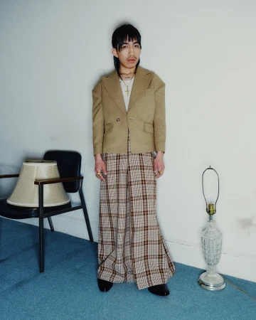 Man wearing a structured tan blazer and plaid trousers standing in an interior space with blue carpet and lamps.