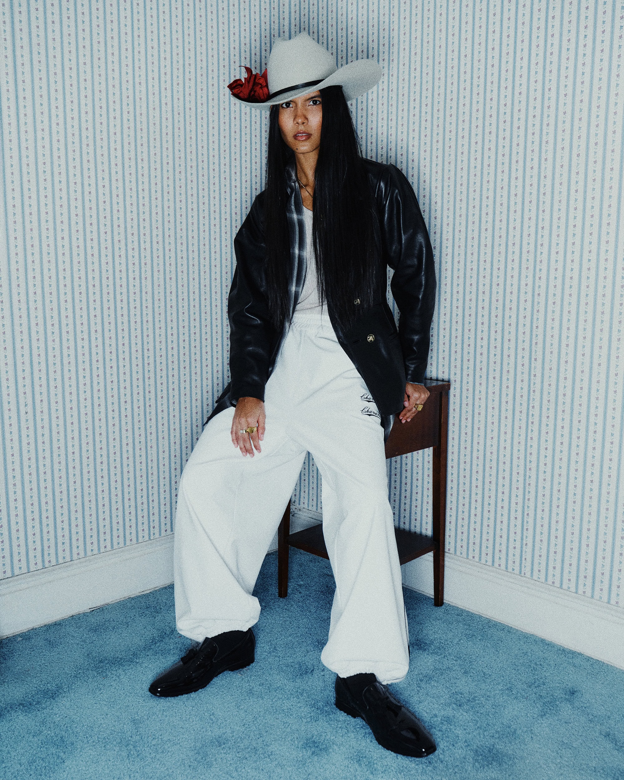 Model wearing white trousers, black jacket, and cowboy hat sitting against blue-striped wallpaper for Willy Chavarria.