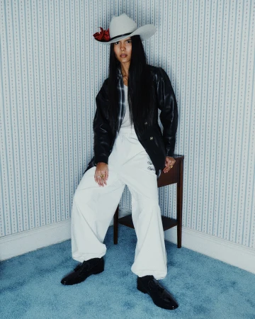 Model wearing white trousers, black jacket, and cowboy hat sitting against blue-striped wallpaper for Willy Chavarria.