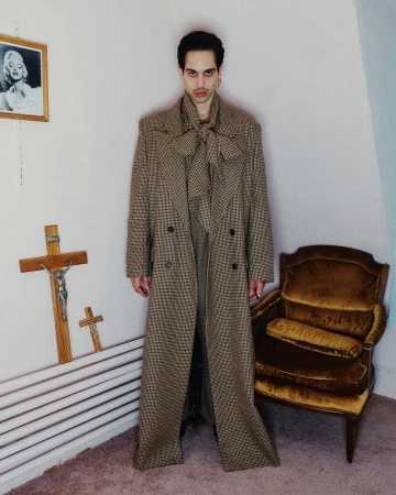 Man wearing a long patterned double-breasted coat standing indoors near a wooden armchair and wall crucifixes.