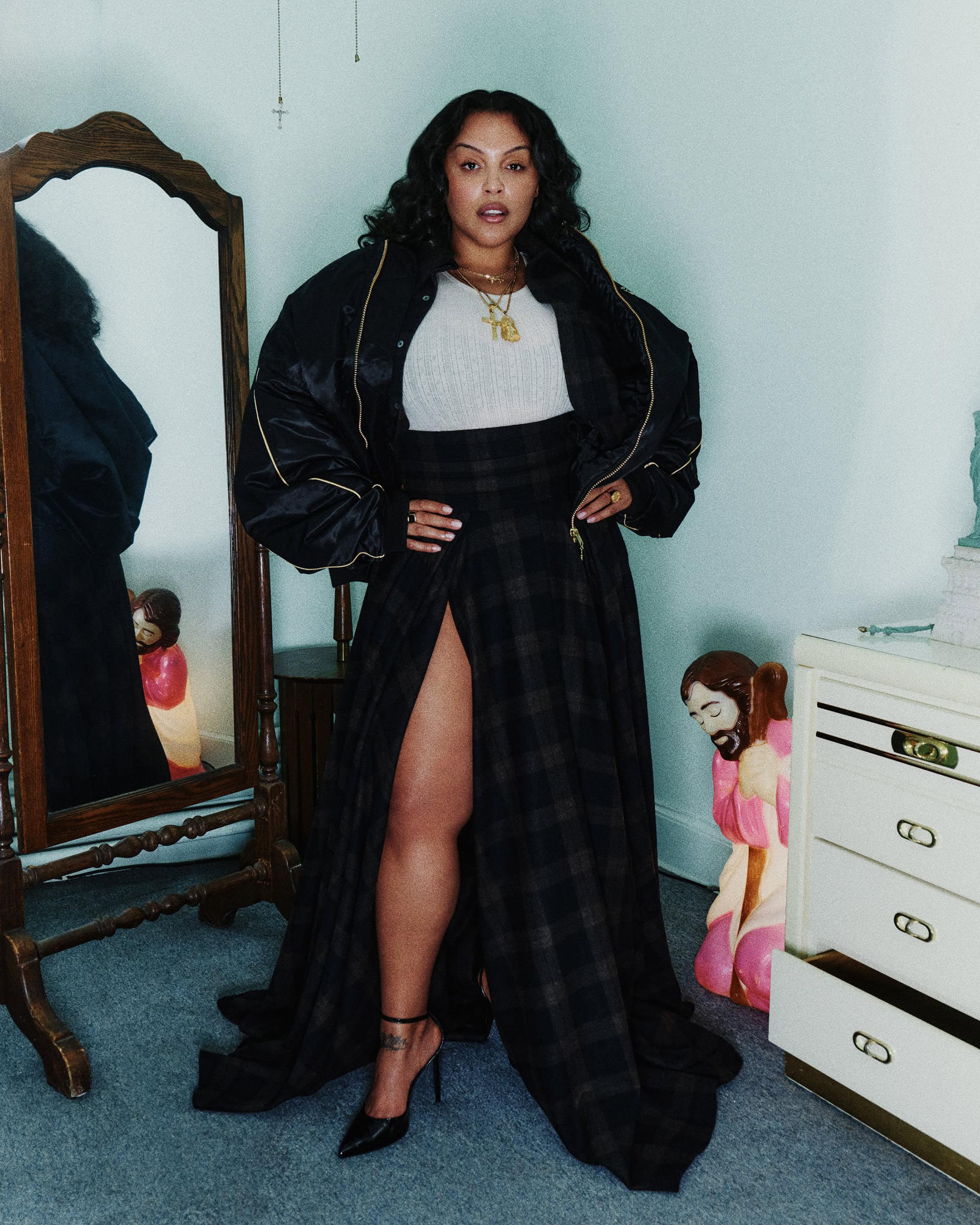 Paloma Elsesser in a black oversized bomber jacket and plaid maxi skirt standing in a domestic bedroom