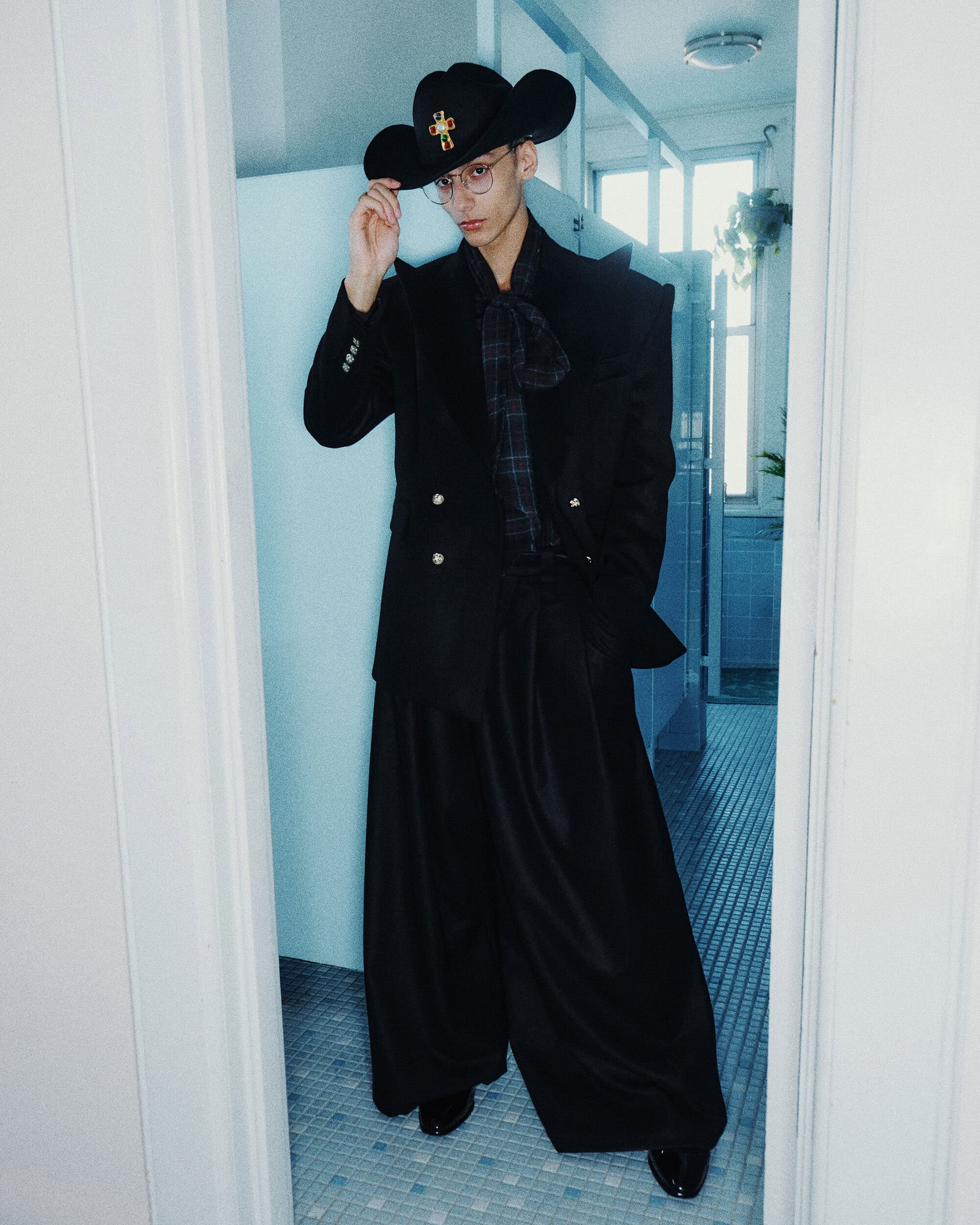 Man wearing a black cowboy hat, structured black blazer, and wide trousers standing in a tiled interior doorway.