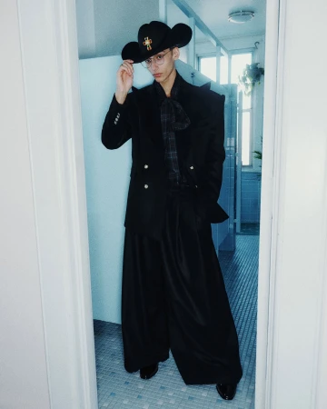 Man wearing a black cowboy hat, structured black blazer, and wide trousers standing in a tiled interior doorway.