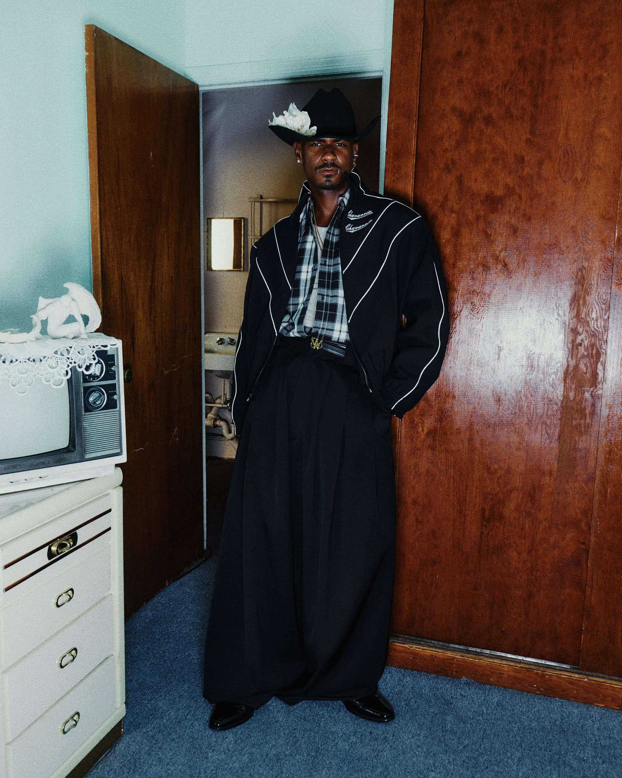 Man wearing a black cowboy hat with white flowers, dark jacket with white piping, and plaid shirt standing in a room.
