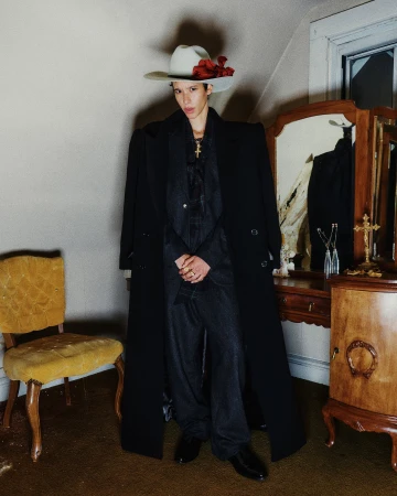 Model wearing a wide-brimmed hat with a red flower, black overcoat, and dark suit standing indoors by a vintage yellow chair and dresser.