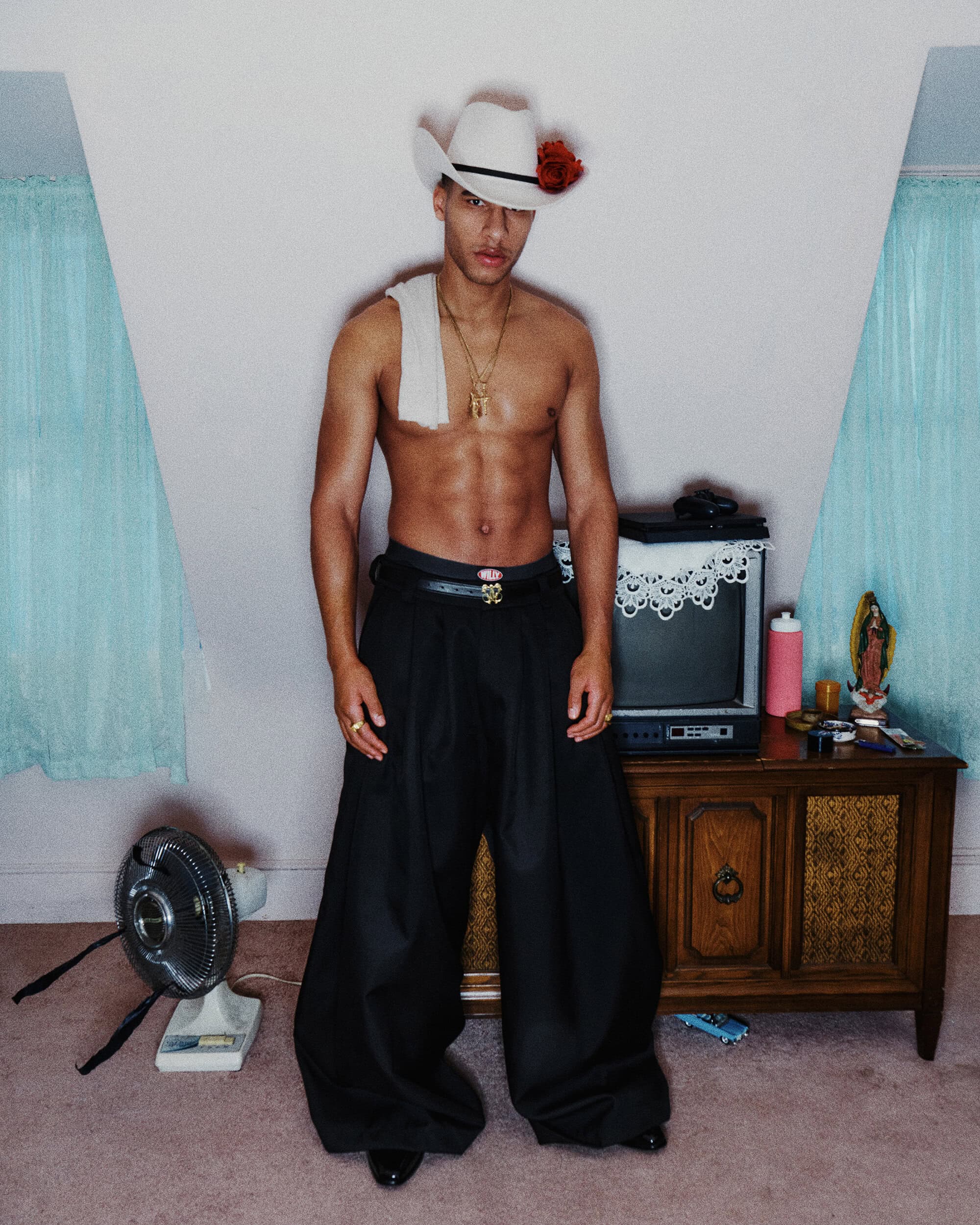 Shirtless man wearing a white cowboy hat with red rose and black wide-leg trousers standing indoors