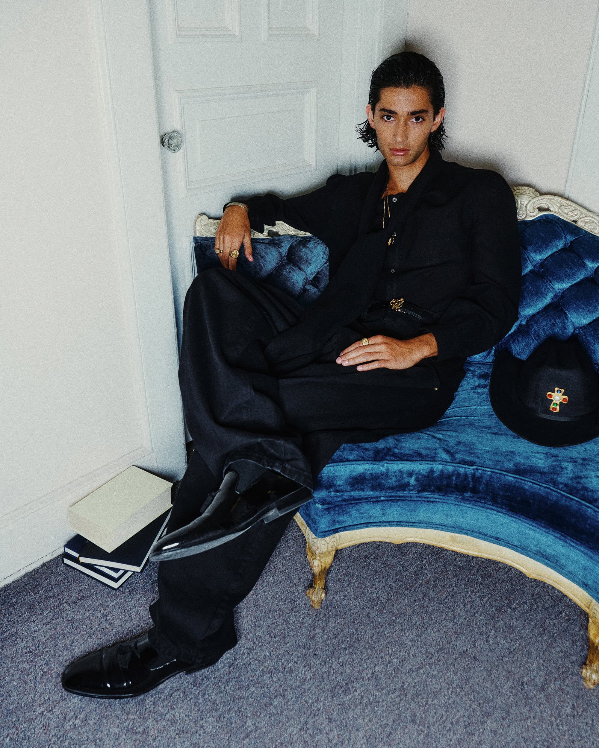 A man seated on a blue velvet couch wearing a black suit and shiny black shoes in a domestic room interior.