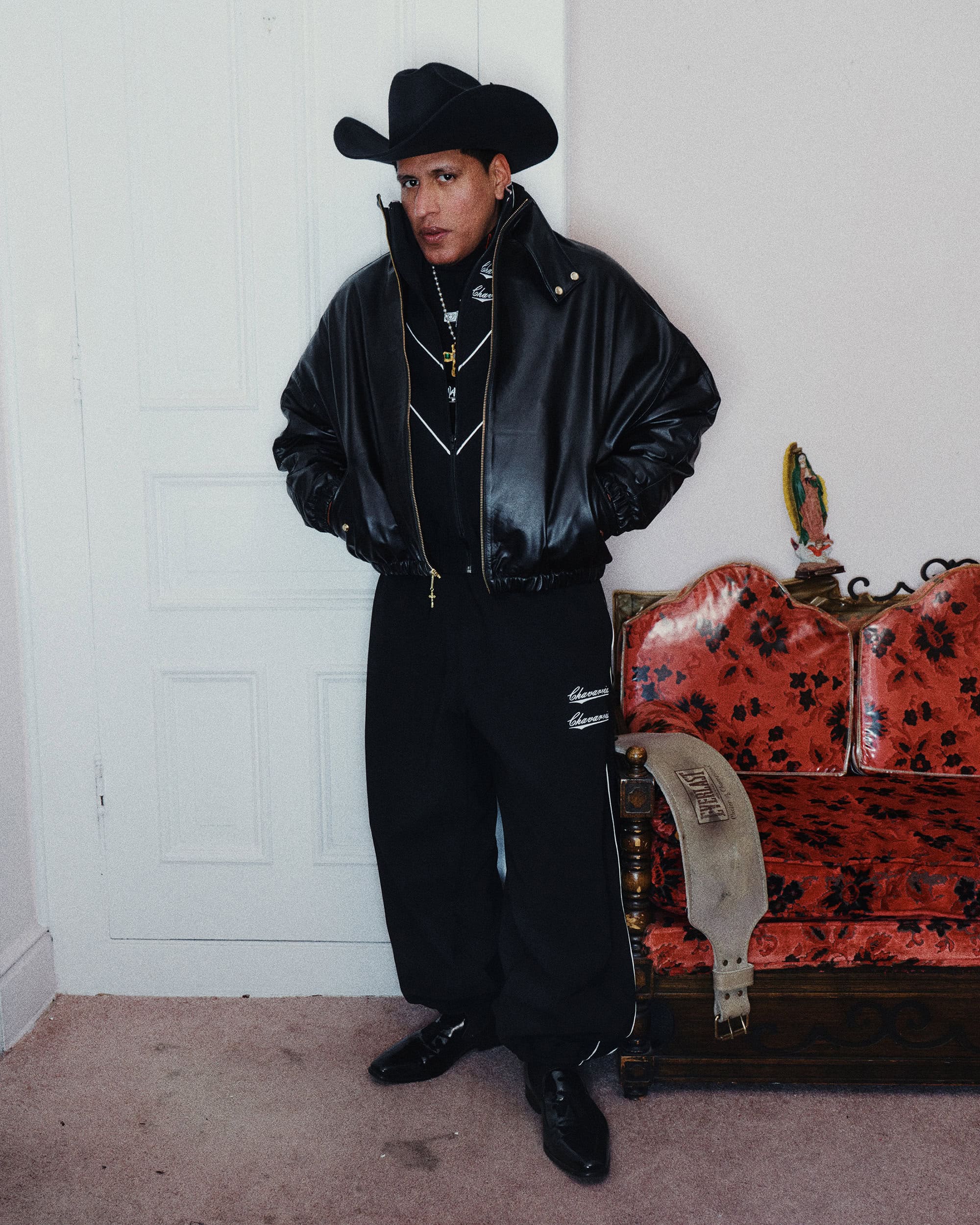 Man wearing a black leather jacket, cowboy hat, and dark trousers standing indoors beside a red patterned sofa.