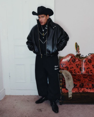 Man wearing a black leather jacket, cowboy hat, and dark trousers standing indoors beside a red patterned sofa.