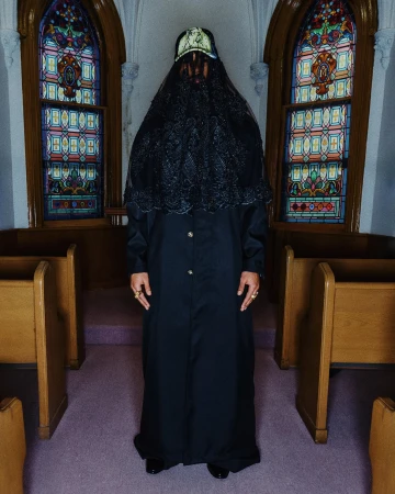 Figure standing centrally in a church interior wearing a long black coat, veiled headwear, and a baseball cap.