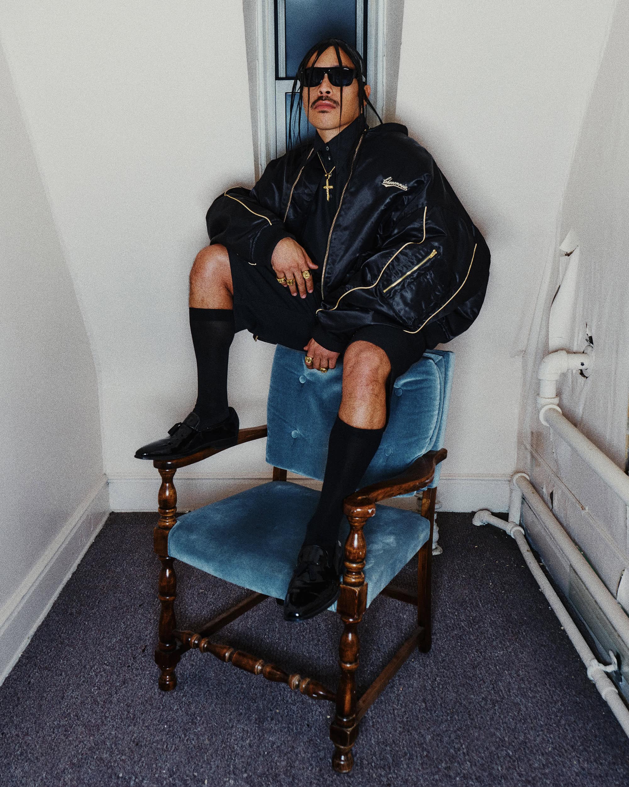 Man wearing a black bomber jacket and shorts seated on a blue velvet chair indoors