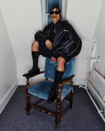 Man wearing a black bomber jacket and shorts seated on a blue velvet chair indoors