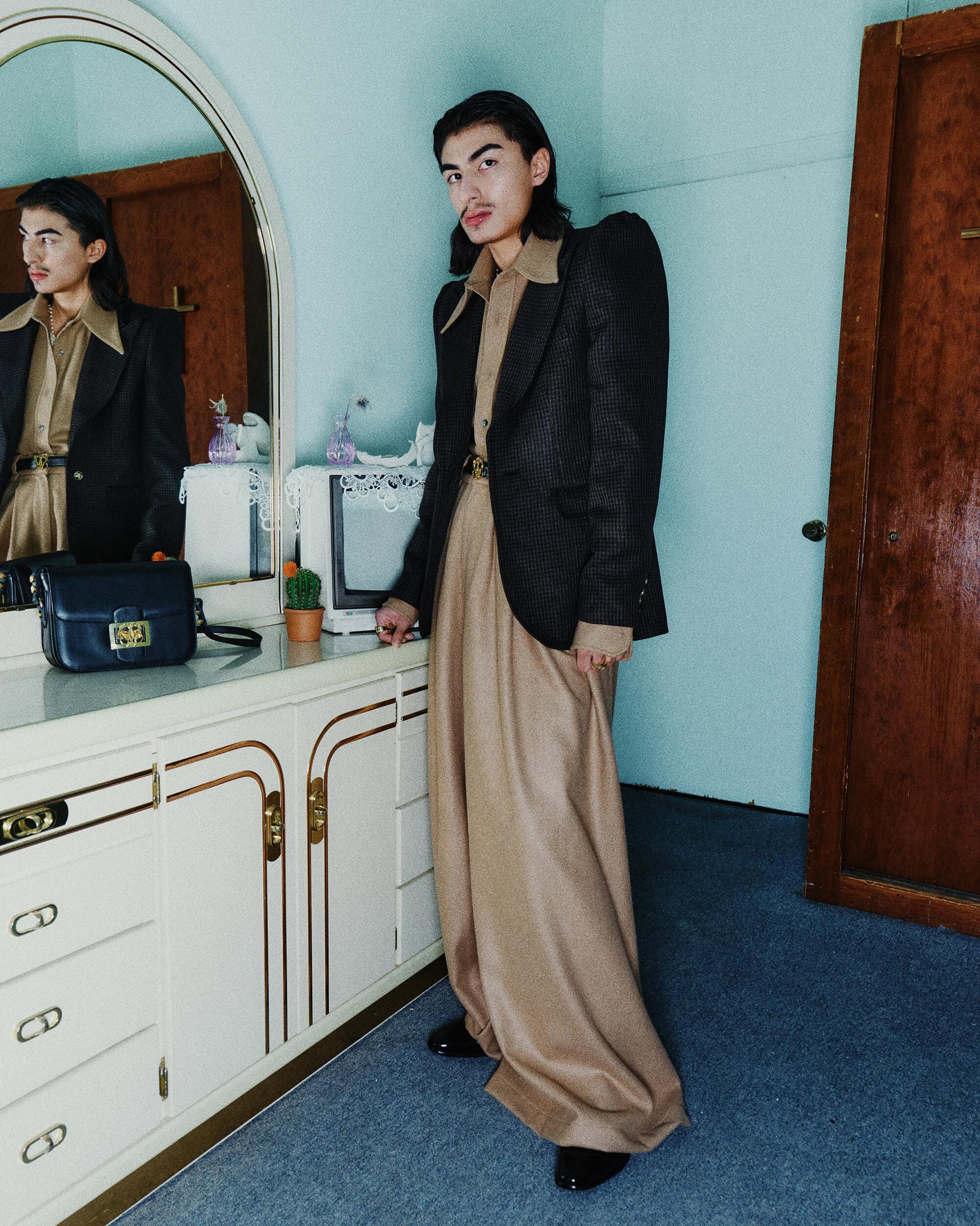 Man wearing a brown collared shirt and structured dark blazer standing in a room with a vanity mirror