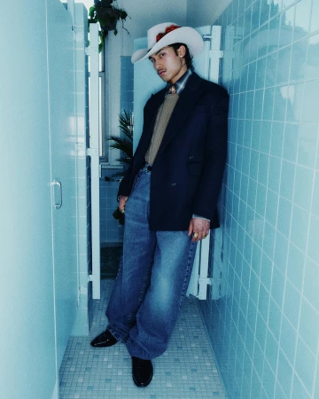 Man wearing a cowboy hat, dark blazer, and wide jeans leaning against a blue tiled wall for Willy Chavarria Fall 2024 campaign.