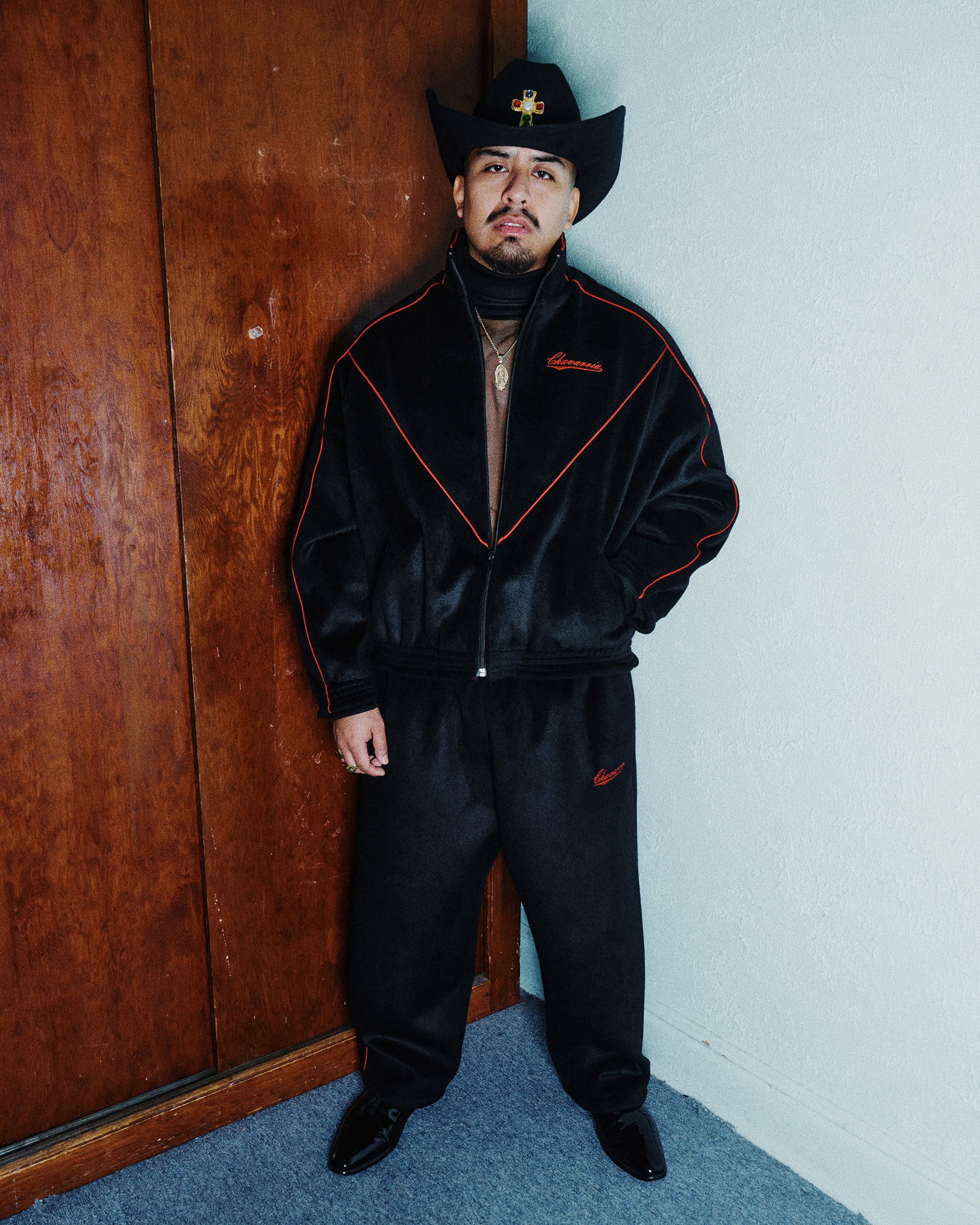 Man wearing a black cowboy hat and matching black track jacket and trousers standing indoors