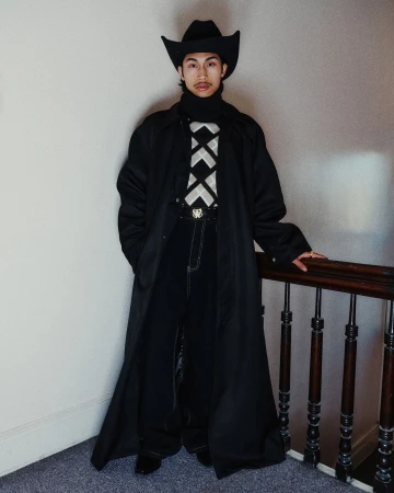 Model wearing a black cowboy hat and long black coat standing beside a dark wooden staircase railing indoors.