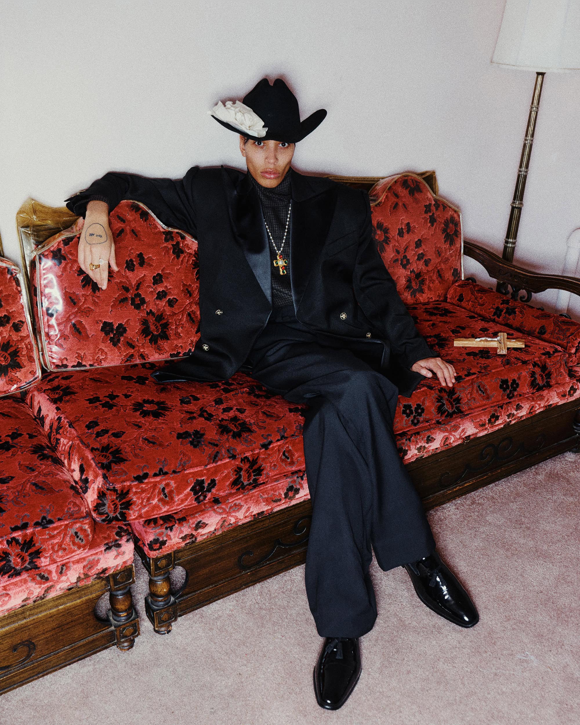 Model seated on a patterned red velvet sofa wearing a black suit and cowboy hat adorned with a white flower.