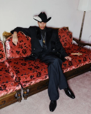 Model seated on a patterned red velvet sofa wearing a black suit and cowboy hat adorned with a white flower.