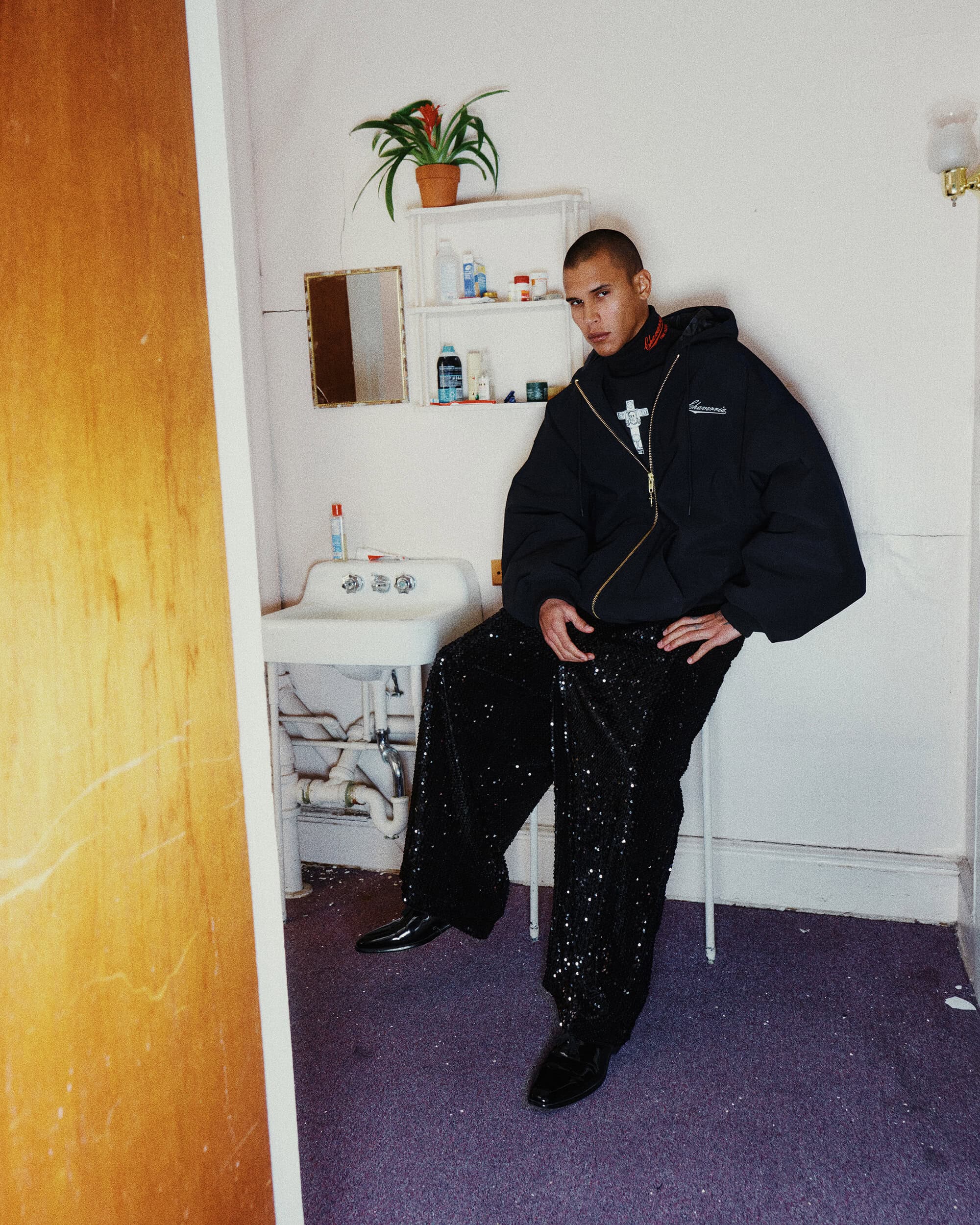 Model wearing a black oversized zip-up hoodie and sequined trousers seated in a bathroom setting.
