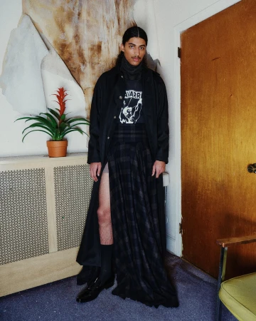 Man wearing a long black coat and plaid skirt standing indoors near a wooden door and potted plant for Willy Chavarria.