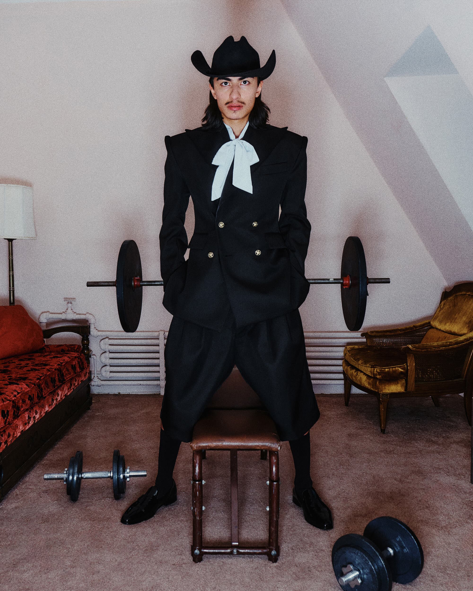 Man wearing a black suit, cowboy hat, and white ascot standing in a room with barbells and exercise weights.