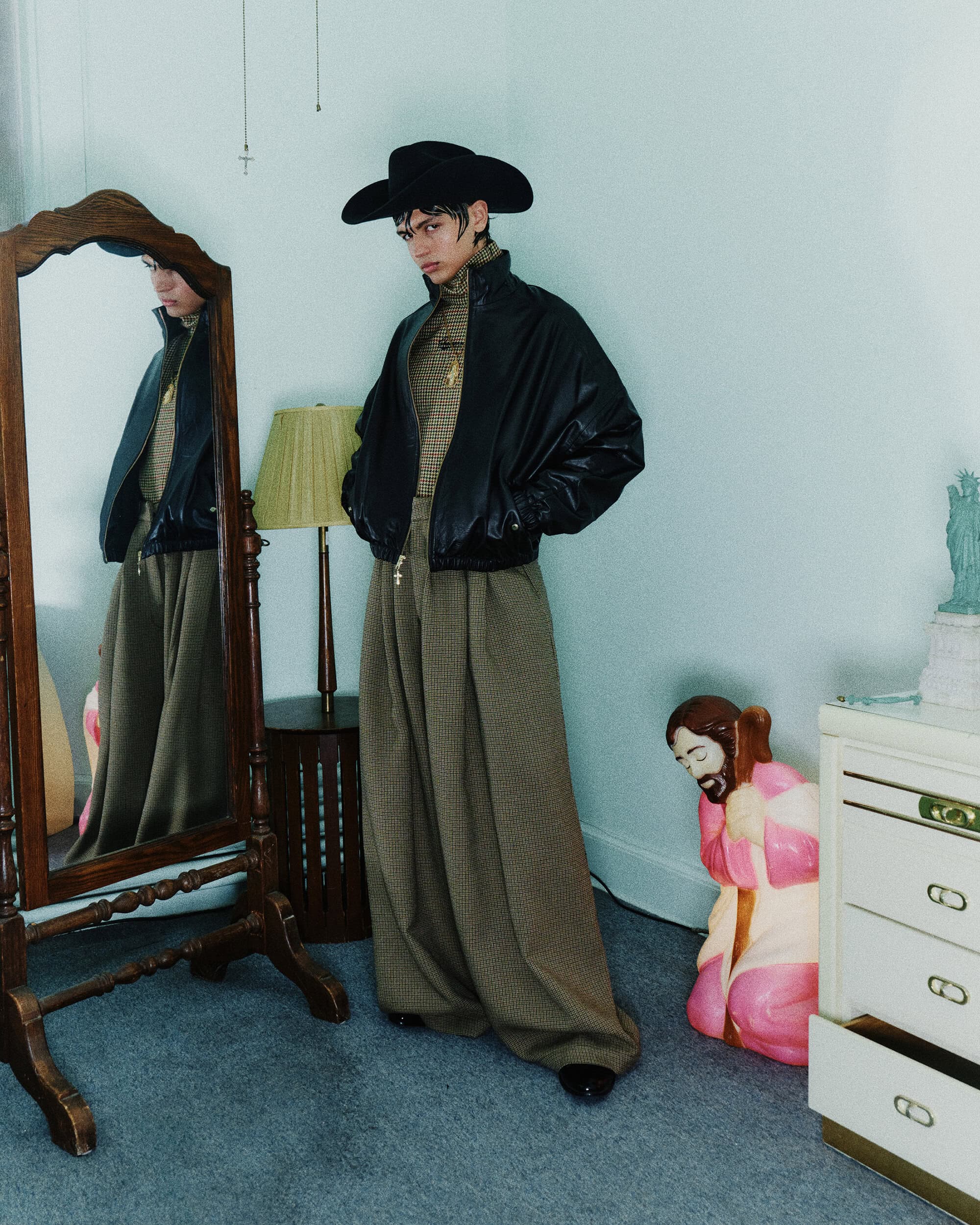 Model wearing a black cowboy hat, black leather jacket, and patterned trousers standing inside a domestic room with a mirror and lamp.