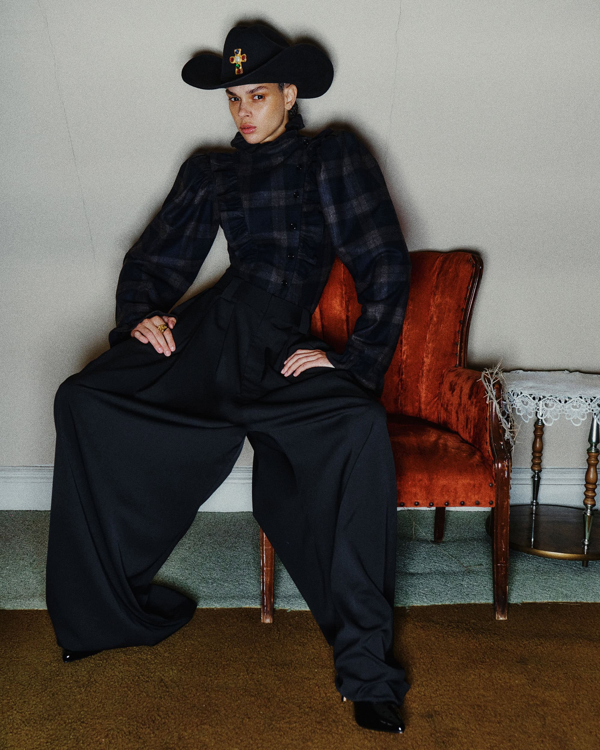 Model wearing a black cowboy hat and plaid top standing with wide black trousers next to an orange chair