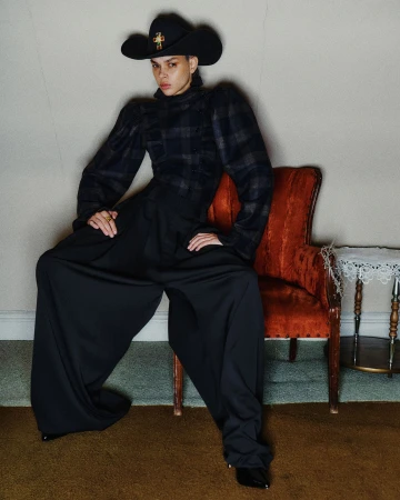Model wearing a black cowboy hat and plaid top standing with wide black trousers next to an orange chair