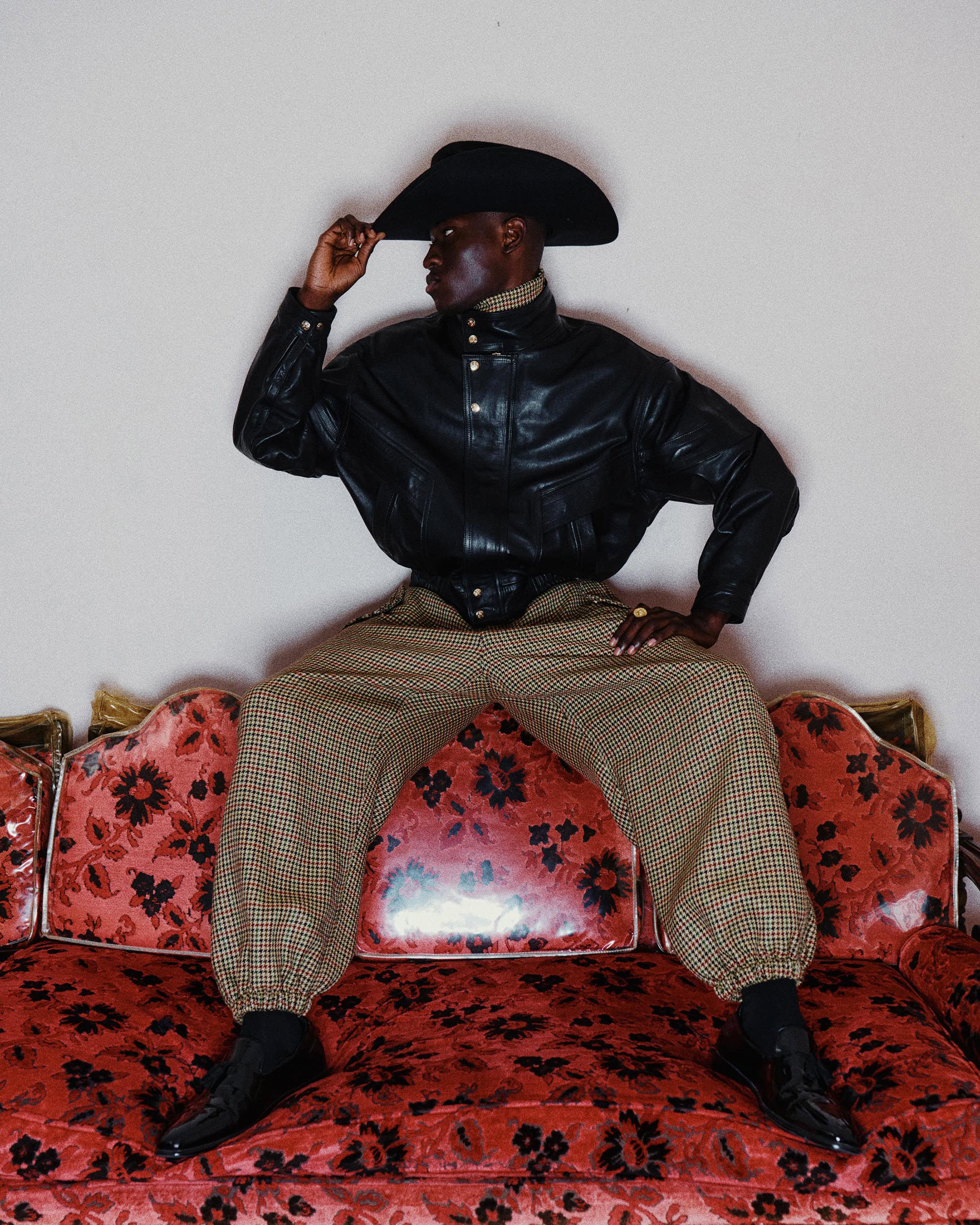 Model wearing a black cowboy hat and leather jacket seated on a red floral sofa for Willy Chavarria Fall 2024 ad campaign.