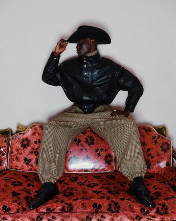 Model wearing a black cowboy hat and leather jacket seated on a red floral sofa for Willy Chavarria Fall 2024 ad campaign.