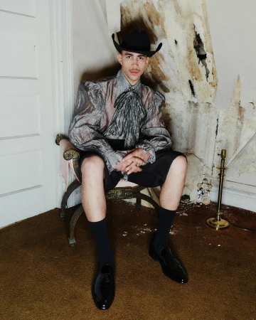Man wearing a cowboy hat and sheer ruffled shirt seated on an antique chair against a distressed wall.