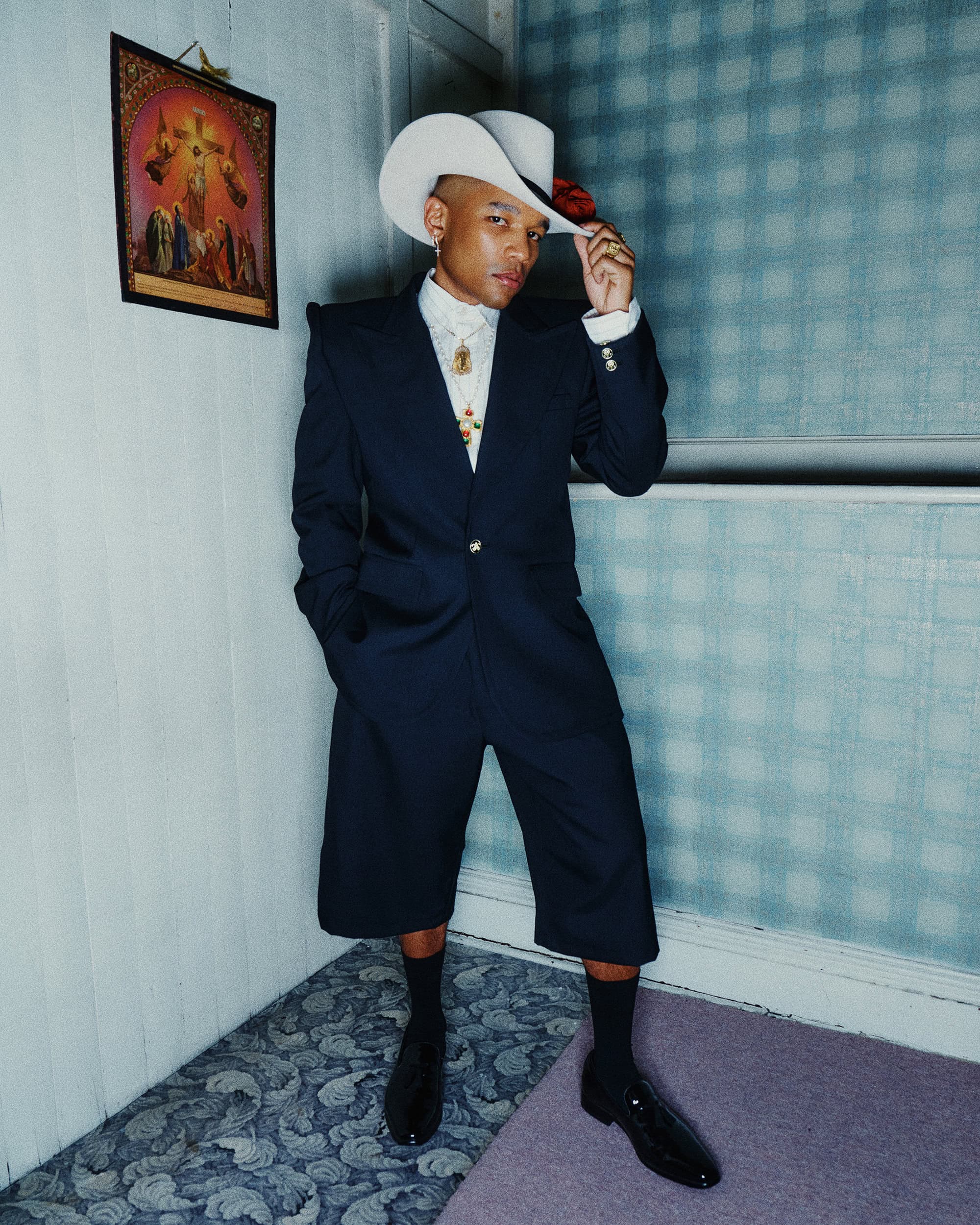 Man wearing a dark suit with shorts, white cowboy hat and black shoes standing against a patterned wall for Willy Chavarria campaign
