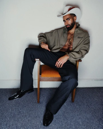 Man with beard wearing white cowboy hat, patterned jacket, and black trousers seated on wooden chair against white wall.