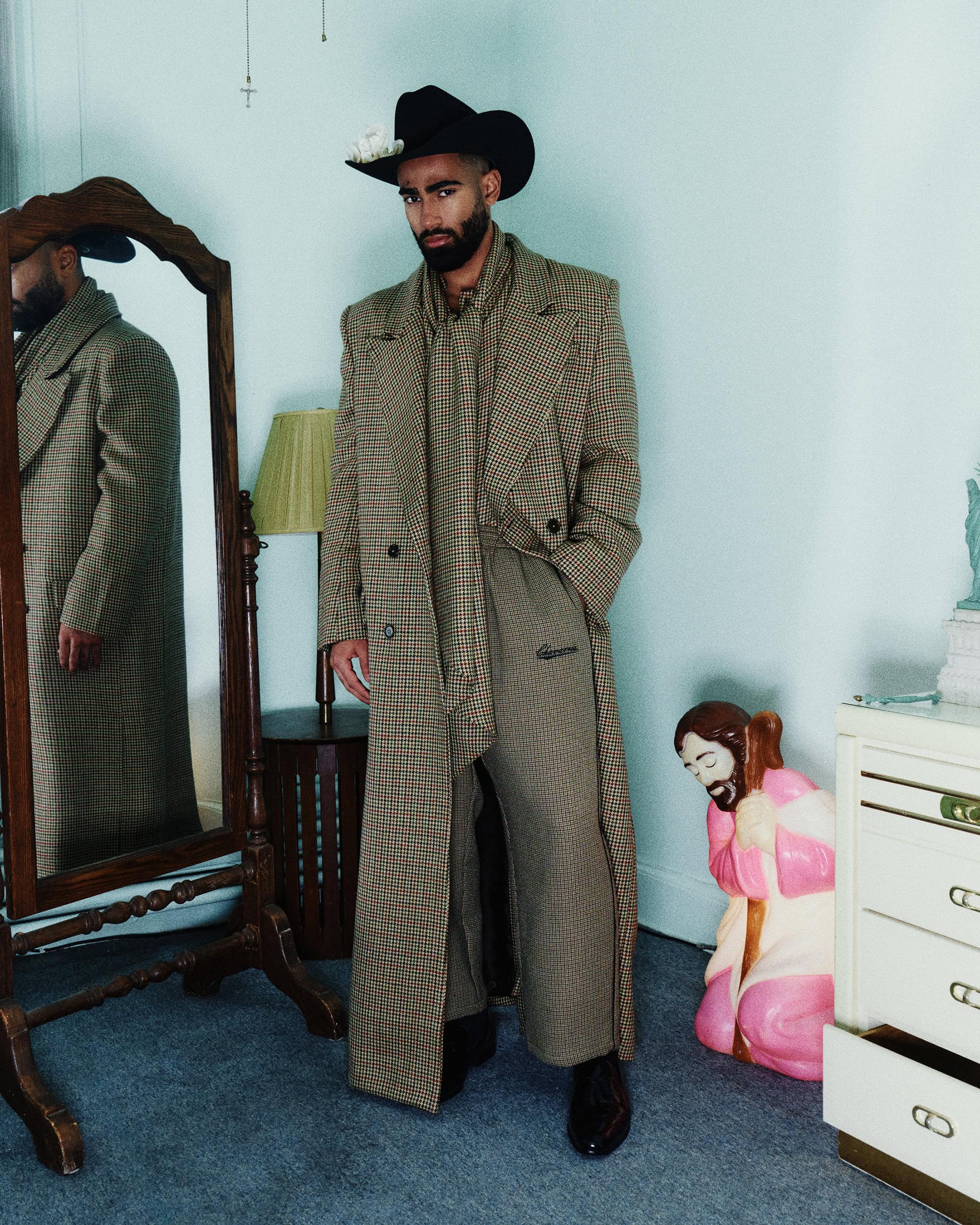 Man wearing a houndstooth coat, matching trousers, and a black cowboy hat standing in a domestic room with a mirror and figurine.