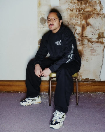 Man seated on a stool wearing a black long-sleeve sweatshirt, loose black pants, and athletic sneakers against a distressed wall.