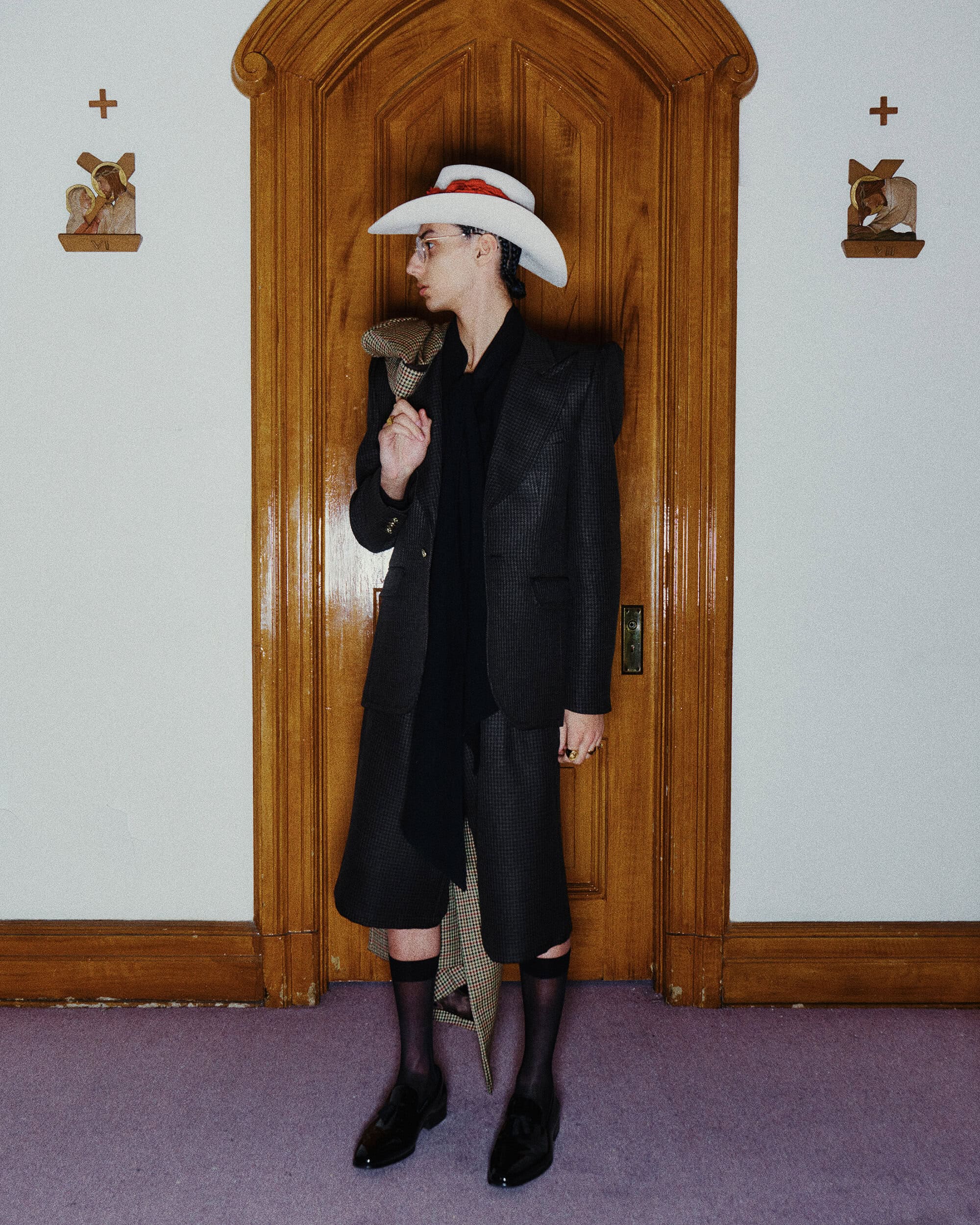 Model wearing a structured dark blazer, shorts, cowboy hat, and sheer black socks standing in front of a wooden doorway.
