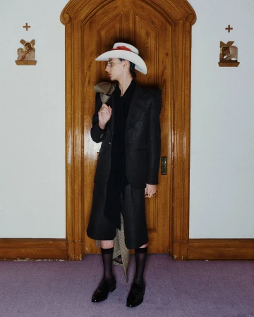 Model wearing a structured dark blazer, shorts, cowboy hat, and sheer black socks standing in front of a wooden doorway.