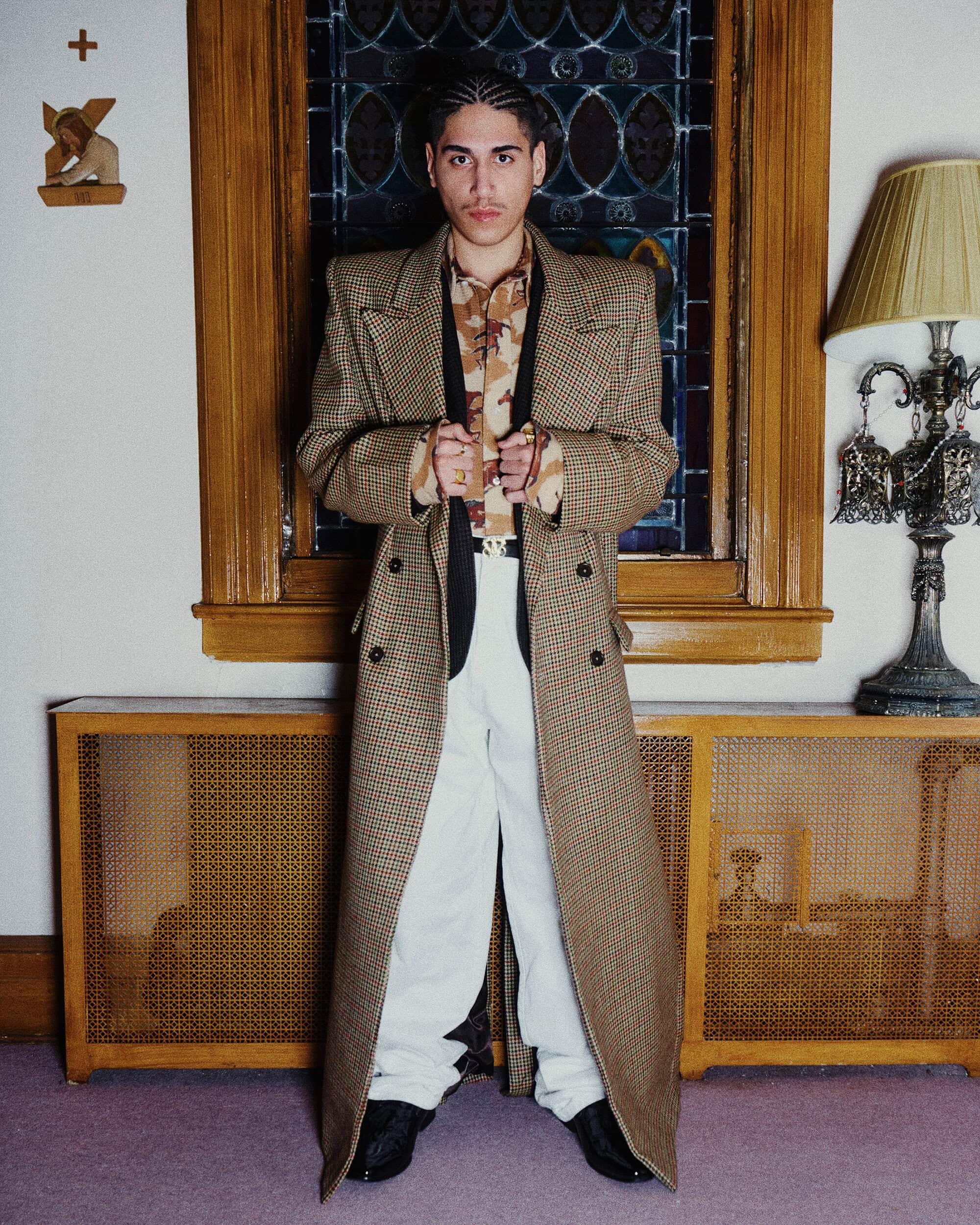 Man wearing a brown houndstooth trench coat and white trousers standing indoors with a stained glass window behind him