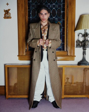 Man wearing a brown houndstooth trench coat and white trousers standing indoors with a stained glass window behind him