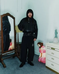 A person with long dark hair wearing a black leather jacket and black trousers stands in a domestic interior with a mirror and dresser.