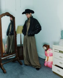 Model wearing a black cowboy hat, black leather jacket, and patterned trousers standing inside a domestic room with a mirror and lamp.