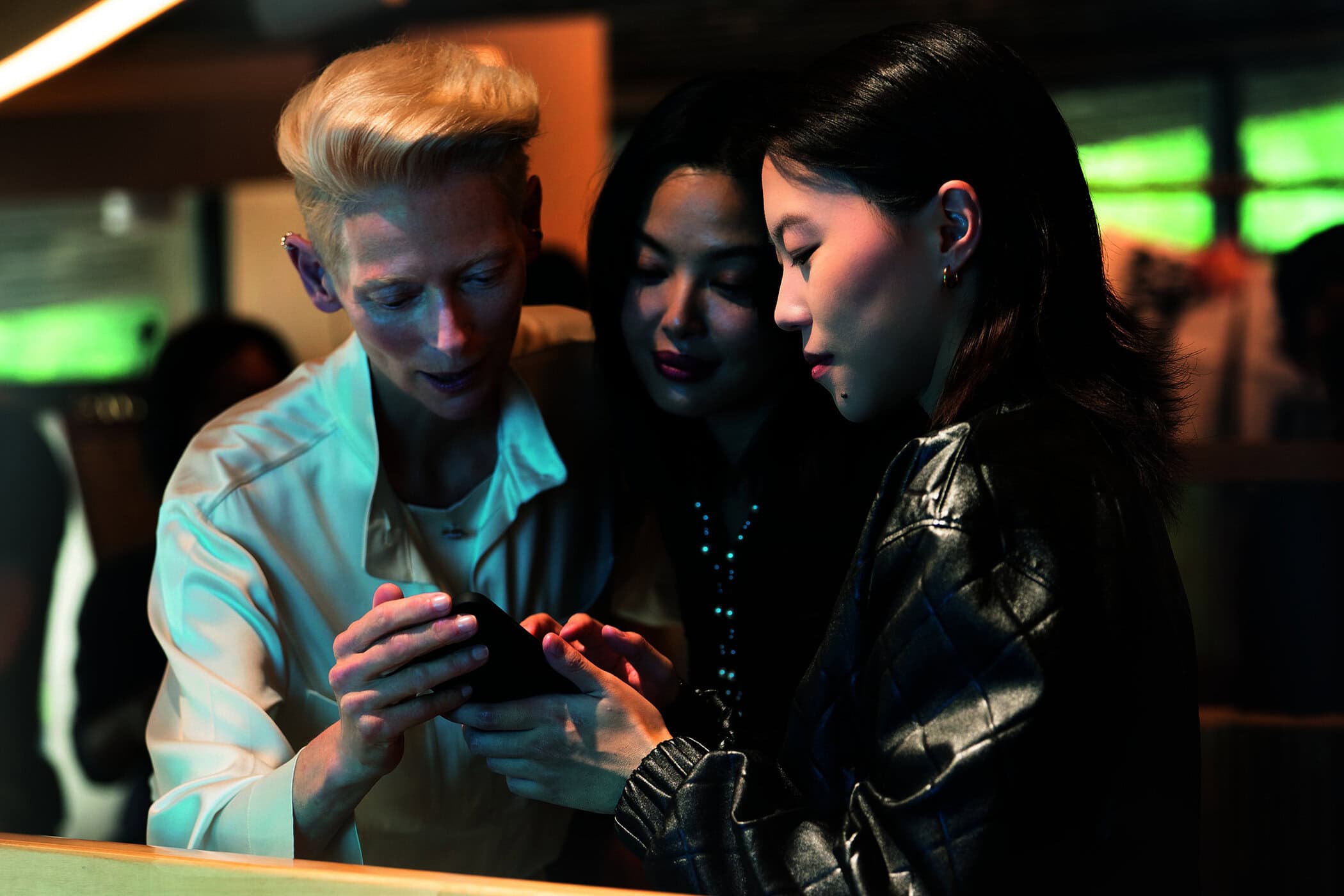 Tilda Swinton and two other women gathered around a smartphone looking down intently in a dimly lit interior setting.